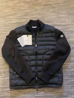 Svart Moncler cardigan - Säljer en snygg cardigan från moncler! Cardiganen är i mycket bra skick. De enda defekterna är att lappen vid nacken sitter lite löst och att cardiganen är lite solblekt på vissa ställen. Nfc-scan funkar på loggan vid armen och qr-koden går att scanna. Allting på bilderna tillkommer! Om ni har några frågor så är det bara att höra av er😊