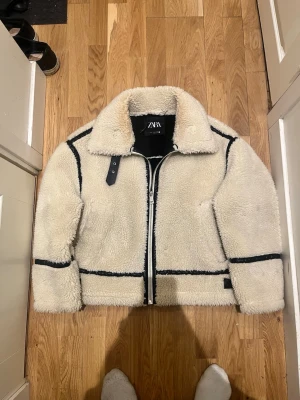 Beige teddyjacka från Zara, M - Säljer en beige teddyjacka från Zara i storlek M. Jackan har svart kontrastsömmar, dragkedja framtill och stor krage. Utsidan är fluffig och mjuk medan insidan är svart och slät. Perfekt för kalla dagar och riktigt snygg till jeans.