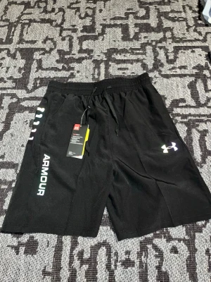 Svarta träningsshorts Under Armour - Snygga svarta träningsshorts från Under Armour med elastisk midja och snörning. Shortsen har logga och text på vänster ben samt coola reflex vita  och färgade detaljer längs sidorna. Tillverkade i lätt och snabbtorkande polyester, perfekta för gymmet eller löprundan.