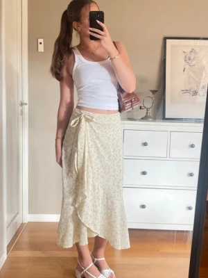 Blommig omlottkjol med volang - Supersöt lång omlottkjol i ljusgul nyans med små blommor i rosa och grönt. Kjolen har en volangkant nertill och knyts i midjan med en rosett. Perfekt för en somrig och fräsch look.