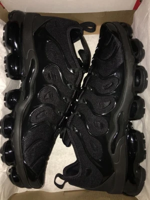 Nike Air Vapormax svarta sneakers - Säljer ett par helsvarta Nike Air Vapormax sneakers med futuristisk design och synliga luftbubblor i sulan. Ovandelen är i mesh och syntet med coola vågformade detaljer och snörning. Perfekta för dig som vill ha en sportig och modern look.
