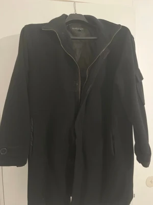 Svart ullkappa med dragkedja - Säljer en svart lång ullkappa från . Kappan har dragkedja framtill, fickor på sidorna och en extra ficka på ärmen. Klassisk krage och knappdetaljer vid ärmsluten. Perfekt för kalla dagar och stilren look.