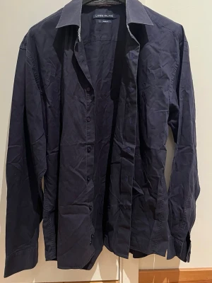 Mörkblå skjorta från Long Island - Snygg mörkblå skjorta från Long Island i 100% bomull. Klassisk modell med lång ärm, knappar framtill och rutigtk mönster på insidan av kragen. Perfekt för dig som gillar stilrena plagg med en liten twist.