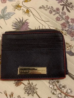 Svart plånbok från Tommy Hilfiger - Snygg svart plånbok från Tommy Hilfiger med guldfärgad logga framtill och röda kanter. Plånboken har dragkedja runtom och flera fack för kort och mynt. Perfekt storlek för att ha med sig överallt och stilren design som passar till allt.