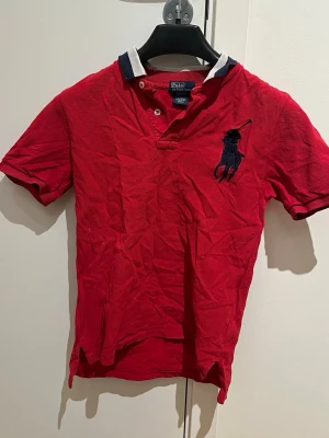 Röd pikétröja från Ralph Lauren - Klassisk röd pikétröja från Ralph Lauren med stor mörkblå broderad logga på bröstet. Tröjan har vit och marinblå krage, korta ärmar och knappar vid halsen. Tillverkad i 100% bomull för en skön och luftig känsla. Barnstorlek
