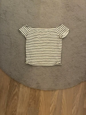 Randig offshoulder topp från H&M - Säljer en vit/lite beige offshoulder topp från H&M med svarta horisontella ränder. Toppen har korta ärmar och en avslappnad passform. Perfekt till sommaren och enkel att matcha med jeans eller kjol.