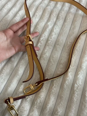 Axelrem - väskband - Säljer en beige axelrem med guldfärgade metalldetaljer och spänne. Remmen har justerbar längd och snygga gula sömmar längs kanterna. Oanvänd! Perfekt för att byta ut eller uppgradera din väska. Inspirerad. ❣️ Passa på - gåva vid varje köp!