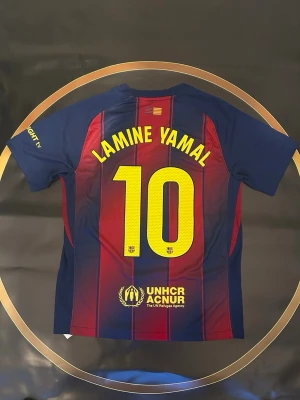 Barça matchtröja Lamine Yamal #10 Nike - Säljer en FC Barcelona matchtröja med Lamine Yamal och nummer 10 på ryggen. Tröjan har klassiska blå och röda ränder, gula detaljer och klubbmärke på bröstet. Tillverkad i lätt polyester med Dri-FIT-teknologi. Sponsorer som Spotify och UNHCR tryckta på framsidan.