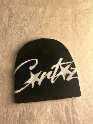 Svart mössa med vit text - Säljer en svart mössa med stor vit text i graffiti-stil över framsidan. Mössan är mjuk och stretchig, perfekt för kyliga dagar. Passar dig som gillar streetwear och vill sticka ut lite extra.