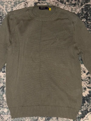 Olivgrön stickad tröja Polo Ralph Lauren - Säljer en olivgrön stickad tröja från Polo Ralph Lauren med klassisk rund hals och diskret broderad logga på bröstet. Tröjan har ribbade muddar vid ärmslut och nederkant, perfekt för lager på lager under höst och vinter.