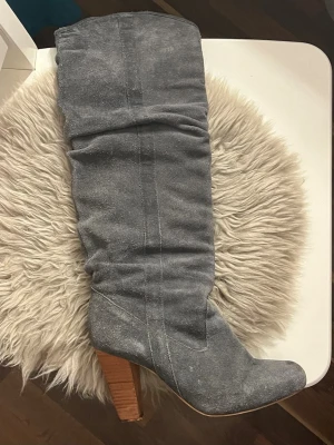 Grå mockastövlar med träklack - Säljer ett par höga stövlar i grå mocka med rund tå och bred träfärgad klack. Skorna har en slouchy passform och är perfekta för dig som vill ha en stilren men ändå trendig look. Klackhöjd 8-9 cm. Längd 51cm