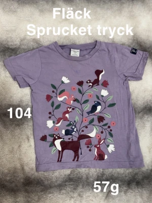 Lila t-shirt med djurmotiv - Söt lila t-shirt från Polarn O. Pyret med korta ärmar och ett färgglatt tryck av skogsdjur och blommor på framsidan.  perfekt för dig som gillar lekfulla motiv.🌸 Fläck samt sprucket tryck. Storlek 104