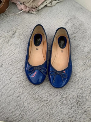 Blå ballerinaskor med rosett - Snygga blå ballerinaskor i lack med rund tå och dekorativ rosett framtill. Skorna har en platt sula och en klassisk, enkel design som passar till många olika outfits. Insidan är beige och de är lätta att bara hoppa i när du ska iväg. De är använda men fortfarande bra skick!