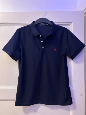 Marinblå pikétröja från Ralph Lauren - Klassisk marinblå pikétröja från Ralph Lauren med korta ärmar och krage. Tröjan har tre vita knappar framtill och den ikoniska röda broderade loggan på bröstet. Tillverkad i bomull för en skön och luftig känsla.