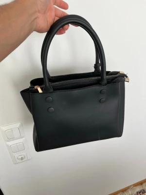 Svart handväska från ECCO i skinn - Stilren svart handväska från ECCO i mjukt skinn med guldfärgade dragkedjor och dubbla handtag. Väskan har tre separata fack med dragkedja och diskreta knappliknande detaljer på framsidan. Perfekt storlek för det viktigaste och snygg att bära till allt.