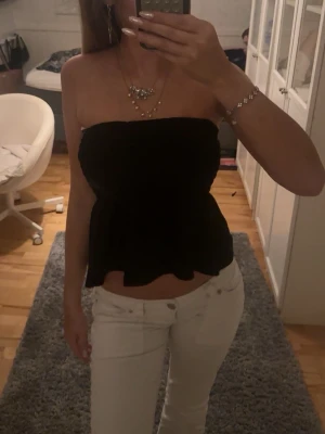 Svart bandeau tubtopp - Säljer en svart bandeau tubtopp med volangkant nertill. Toppen är axelbandslös och har en lätt utsvängd form som ger en snygg siluett. Perfekt att matcha med jeans eller kjol för en trendig look.