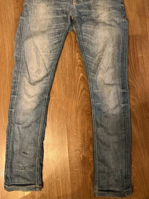 Blå Nudie Jeans, W28 L32 - Säljer ett par klassiska blå jeans från Nudie Jeans med snygg slitning och kontrastsömmar. Modellen har raka ben och normal passform. Tillverkade i slitstarkt bomullstyg och perfekta för dig som gillar en avslappnad men ändå stilren look. De är kapade från L32-L30 W28