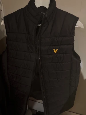 Svart dunväst från Lyle & Scott - Säljer en svart quiltad dunväst från Lyle & Scott med gul logga på bröstet. Västen har hög krage och dragkedja framtill. Perfekt att slänga över en hoodie eller tröja för extra värme. Enkel och stilren design som funkar till det mesta.