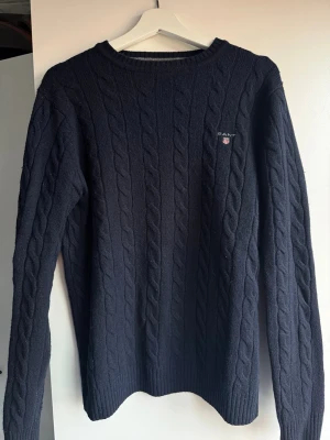 Mörkblå kabelstickad tröja från GANT - Kabelstickad mörkblå tröja från GANT i mjuk lammull. Klassisk rund halsringning och diskret logga på bröstet. Perfekt för dig som gillar stilrena och tidlösa plagg med snygg struktur. Passar till allt och är superenkel att matcha.