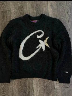 Svart stickad tröja med stjärna från Corteiz - Svart stickad tröja från Corteiz med stor vit C och stjärna på framsidan. Rund hals, ribbade muddar och långärmad. Broderad logga på ärmen. Perfekt för dig som gillar coola detaljer och mjuka material.