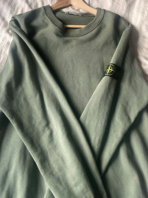 Grön sweatshirt från Stone Island - Säljer en grön sweatshirt från Stone Island med klassisk rund hals och ikonisk patch på ärmen. Tröjan har en relaxed passform och är tillverkad i mjuk bomull, perfekt för en clean och stilren look.