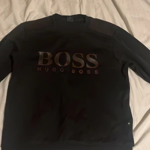 Svart sweatshirt från Hugo Boss - Säljer en svart sweatshirt från Hugo Boss med stor BOSS-logga i relief på bröstet. Tröjan har rund halsringning och långa ärmar. Materialet känns mjukt och har en clean look, perfekt för dig som gillar stilrena plagg med tydlig branding. Den har också färgen mörk röd på axlarna.