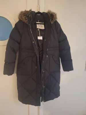 Svart lång dunjacka Chiara Forthi - Säljer en svart lång dunjacka från Chiara Forthi med huva och fejkpälsdetalj. Jackan har quiltat mönster, dragkedja framtill och stora fickor med lock. Perfekt för kalla dagar och riktigt snygg till vintern.