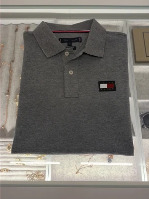 Grå pikétröja Tommy Hilfiger M - Snygg grå pikétröja från Tommy Hilfiger i slim fit med klassisk krage och två knappar. Tröjan har det ikoniska Hilfiger-logotypen broderad på bröstet och är tillverkad i mjuk bomullspiké. Perfekt till jeans eller shorts.