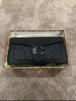 Svart axelväska - Snygg svart axelväska med klassiskt monogrammönster och stor metallspänne framtill. Väskan har avtagbar och justerbar axelrem samt detaljer i skinn och polyester. Perfekt storlek för det viktigaste och levereras med originalbox och dustbag.