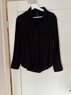 Svart skjorta från New yorker. - Klassisk svart skjorta från New yorker med lång ärm och enkel krage. Skjortan har en rak passform och knappar framtill som är diskret dolda. Perfekt att styla till jeans eller kjol för en clean look.