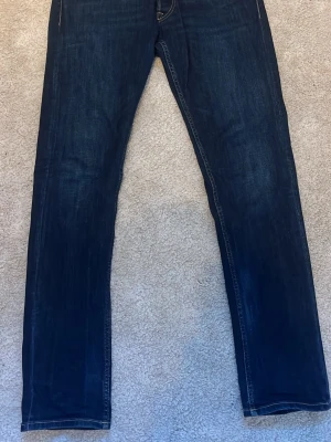 Replay Grover mörkblå jeans - Säljer ett par Replay Grover sopiga mörkblå jeans med klassisk femficksdesign och snygga kontrastsömmar. Jeansen har raka ben och normal passform, tillverkade i slitstarkt denimtyg. Perfekta för dig som gillar stilrena och tidlösa jeans.