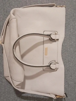 Ljusrosa/lila handväska med gulddetaljer - Stilren ljusrosa/lila handväska i skinnimitation med två rejäla handtag och guldfärgade metalldetaljer. Väskan har en klassisk, fyrkantig form och är perfekt för att bära allt du behöver till vardags. Enkel och snygg design som passar till det mesta. Man kan dock inte överfylla väskan, då åker dragkedjan upp så fyll den måttligt. 