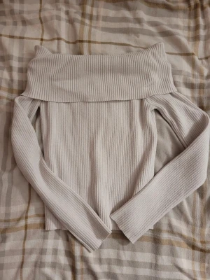 Ljusribbad offshoulder topp - Säljer en ljus, ribbad offshoulder topp med långa ärmar. Toppen har en bred, vikt kant upptill som ger en snygg och trendig look. Perfekt att styla med jeans eller kjol för en enkel men chic outfit.