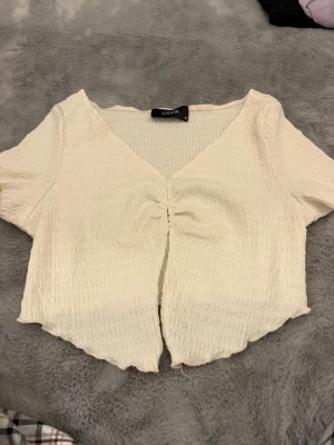 Krämvit croppad topp från Cider - Säljer en krämvit croppad topp från Cider med korta ärmar och v-ringning. Toppen har en ribbad struktur och snygg snörningsdetalj framtill som ger en lätt rynkad effekt. Perfekt till jeans eller kjol för en chill look.