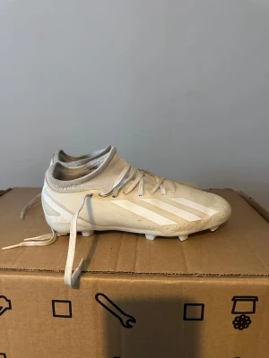 Vita fotbollsskor från Adidas - Säljer ett par vita fotbollsskor från Adidas med klassiska tre ränder på sidan. Skorna har snörning, låg profil och dobbar för bra grepp på planen. Tillverkade i syntetmaterial med stickad kant runt ankeln för extra komfort.