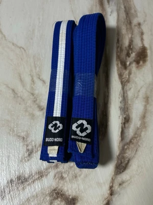 Blått Budo-Nord bälte med vit rand - Säljer ett blått kampsportsbälte från Budo-Nord, tillverkat i 55% bomull och 45% polyester. Ena bältet har en vit rand längs hela längden, medan det andra är helblått. Perfekt för träning inom judo, karate eller liknande.