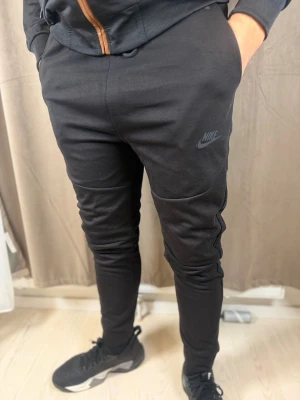 Svarta mjukisbyxor från Nike - Säljer ett par svarta mjukisbyxor från Nike med diskret logga på låret. Byxorna har smal passform, muddar vid bensluten och är tillverkade i mjukt bomullsmaterial. Perfekta för chill eller träning och riktigt sköna att bära.