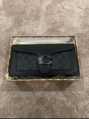 Svart axelväska från Coach - Säljer en svart axelväska från Coach med klassiskt monogrammönster och stor silverfärgad C-detalj på locket. Väskan har avtagbar och justerbar axelrem samt är tillverkad i skinn och polyester. Perfekt storlek för det viktigaste och levereras med originalbox och dustbag.