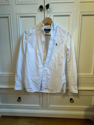 Vit skjorta Polo Ralph Lauren slim fit - Klassisk vit skjorta från Polo Ralph Lauren i slim fit-modell. Skjortan har lång ärm, knappar framtill och den ikoniska broderade loggan på bröstet. Tillverkad i mjuk bomull och passar perfekt till en clean och stilren look.