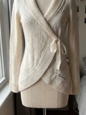 Vit omlottkofta från Bondelid - Säljer en vit stickad omlottkofta från Bondelid i storlek S. Koftan har långa ärmar, ribbade muddar och ett snyggt knytband i midjan. Detaljerad med små hålmönster och broderad Bondelid-logga framtill. Perfekt för lager-på-lager under kyliga dagar.