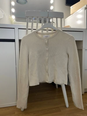 Beige ribbad kofta från Zara - Säljer en beige ribbad kofta från Zara i storlek 152. Koftan har rund hals, långa ärmar och knäppning framtill med matchande knappar. Perfekt att slänga över en topp för en chill och enkel look.