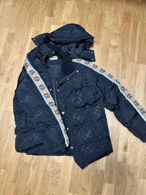 Svart pufferjacka från Gucci XL - Säljer en svart pufferjacka från Gucci i storlek XL. Jackan har ikoniska GG-loggor ton-i-ton över hela tyget och beigea band med GG-mönster längs ärmarna. Utrustad med dragkedja och tryckknappar samt huva. Materialet är polyester och jackan har en riktigt lyxig vibe. Priset är ej 100 satt