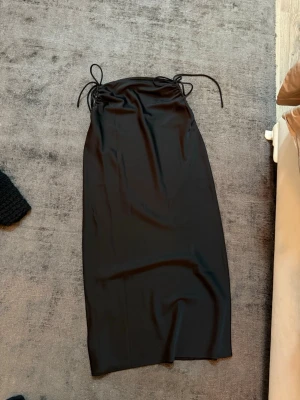 Svart långkjol med snörning - Säljer en svart långkjol i glansigt material med snygga snörningsdetaljer i midjan. Kjolen har en rak och enkel siluett som ger en clean look. Perfekt att styla med crop top eller oversized tröja för en trendig vibe. xs