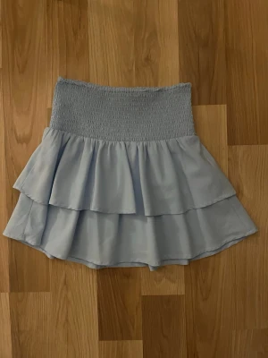 Ljusblå volangkjol från Gina Tricot - Supersöt ljusblå kortkjol från Gina Tricot med bred smock i midjan och dubbla volanger. Kjolen har en flowy känsla och är perfekt för dig som gillar en feminin och trendig stil. Tillverkad i ett mjukt och lätt material som känns skönt mot huden.