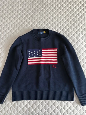 Stickad tröja Polo Ralph Lauren (mörkblå) - Säljer en snygg stickad tröja från Polo Ralph Lauren med amerikansk flagga på, samt RL-broderi. Klassisk rund halsringning och ribbade muddar vid ärmslut och nederkant. Passar bra för en klassisk oldmoney look! 