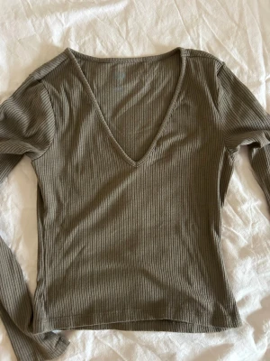 Ribbad khakigrön långärmad topp - Säljer en ribbad långärmad topp i khakigrönt med djup v-ringning. Toppen har en slim passform och är tillverkad i mjukt bomullsmaterial som känns skönt mot huden