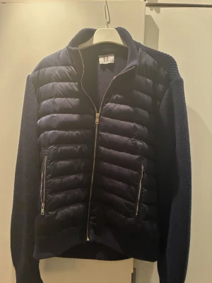 Mörkblå cardigan från Elegance Essentials - Säljer en mörkblå cardigan från Elegance Essentials. Cardiganen har quiltad front i syntetmaterial och stickade ärmar samt rygg. Utrustad med dragkedja framtill och två sidofickor med dragkedja. Perfekt för dig som vill ha en stilren och bekväm jacka. Andvänd en gång säljer den för att den är för stor.