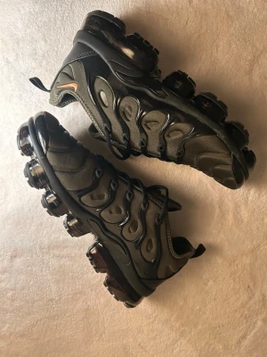 Nike Air Vapormax Plus svart/grön - Nike Air Vapormax Plus i svart och mörkgrön med ikonisk bubblig sula och diskret orange swoosh på sidan. Ovandelen är i syntet och textil med vågiga detaljer och snörning. Snygg och futuristisk sneaker som sticker ut med sin unika design.