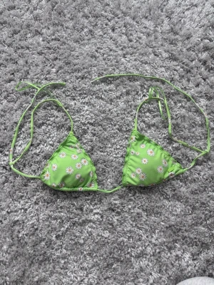 Grön blommig bikiniöverdel med knyt - Snygg grön bikiniöverdel med vita och gula blommor. Triangelformade kupor och tunna knytband i nacken och ryggen för justerbar passform. Perfekt för soliga dagar på stranden.