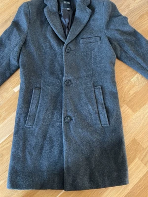 Mörkgrå ullrock från Only & Sons XS - Snygg mörkgrå rock från Only & Sons i storlek XS. Klassisk modell med tre knappar framtill, två fickor och en bröstficka. Tillverkad i mjuk ullmix som ger en stilren och tidlös look. Perfekt för dig som vill ha en clean och enkel jacka.